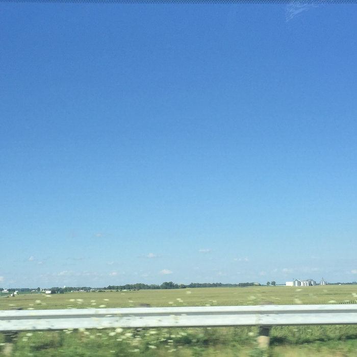 A picture taken from a car on a highway. The metal guardrail is visible. It's a mostly clear, blue sky, with a clear, green field. There's a treeline in the distance.